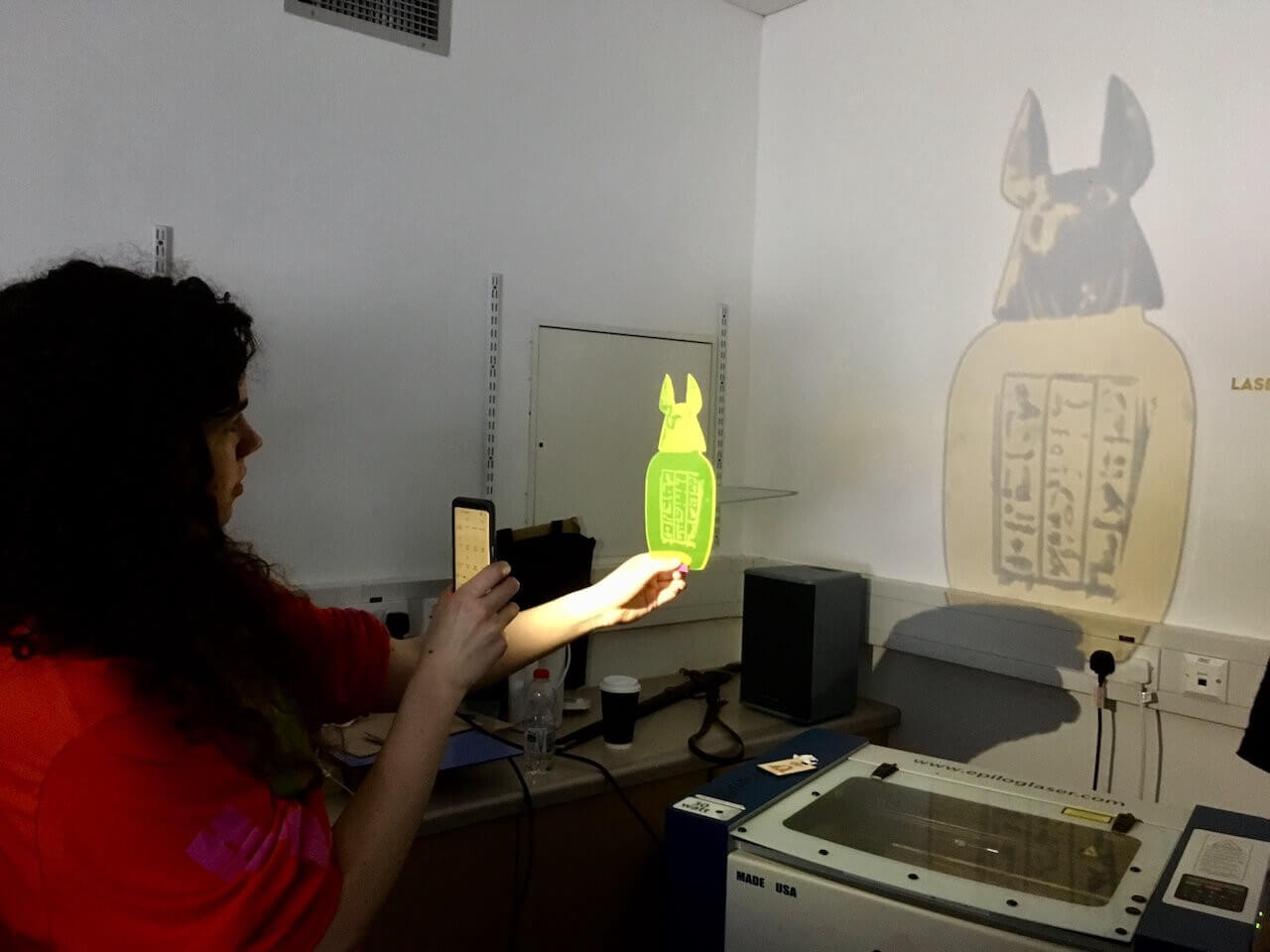 A woman (me) shining a light through a transparent, acrylic canopic jar cutout to project its image on a wall.